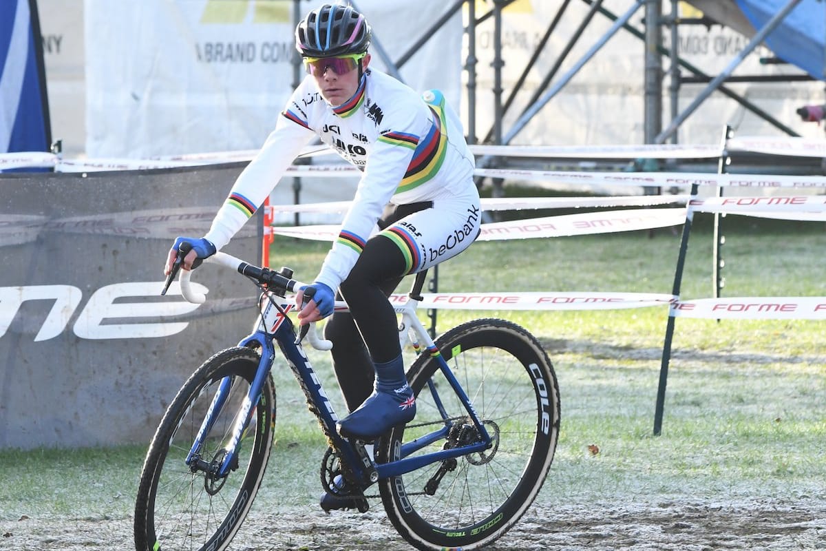 Cyclist  Ben Tulett riding along a muddy field with banners and scaffolding in the background