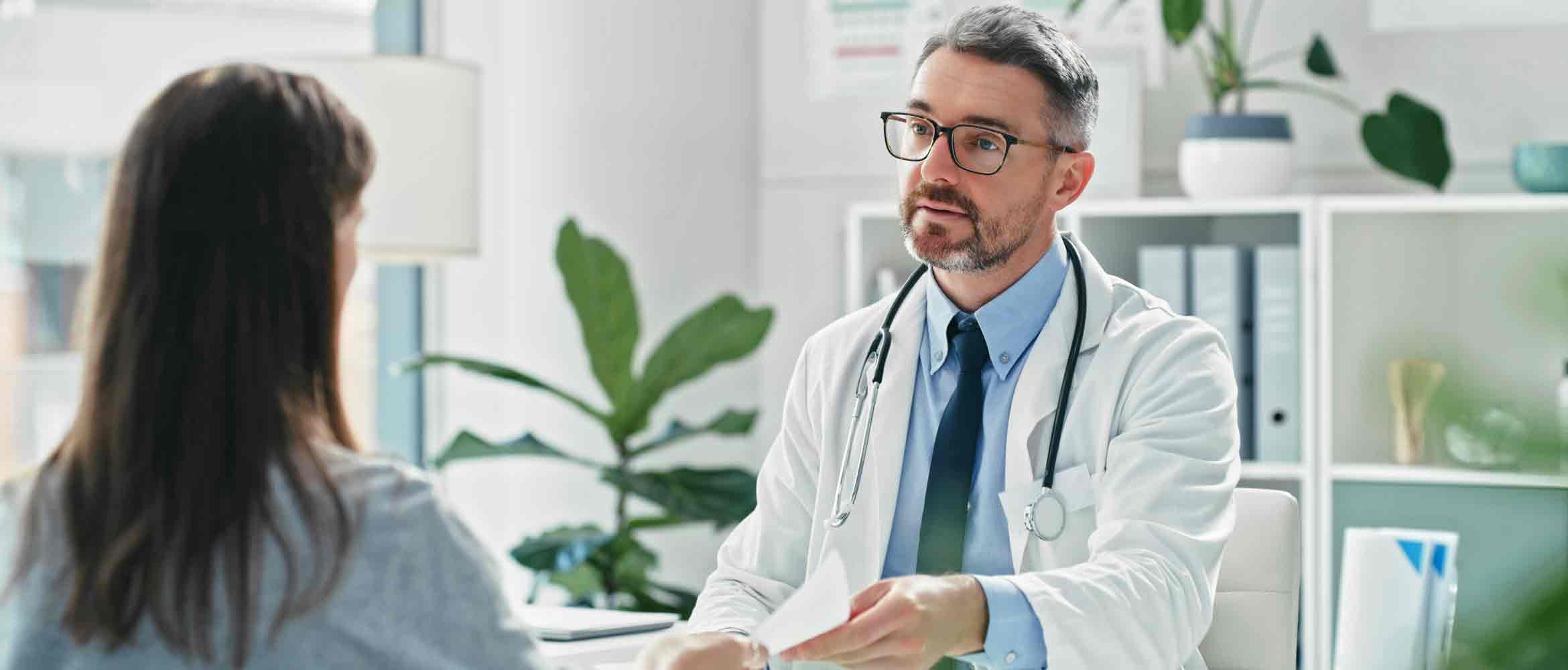 Male doctor handing over a prescription at a desk to a female patient in a doctor's sur