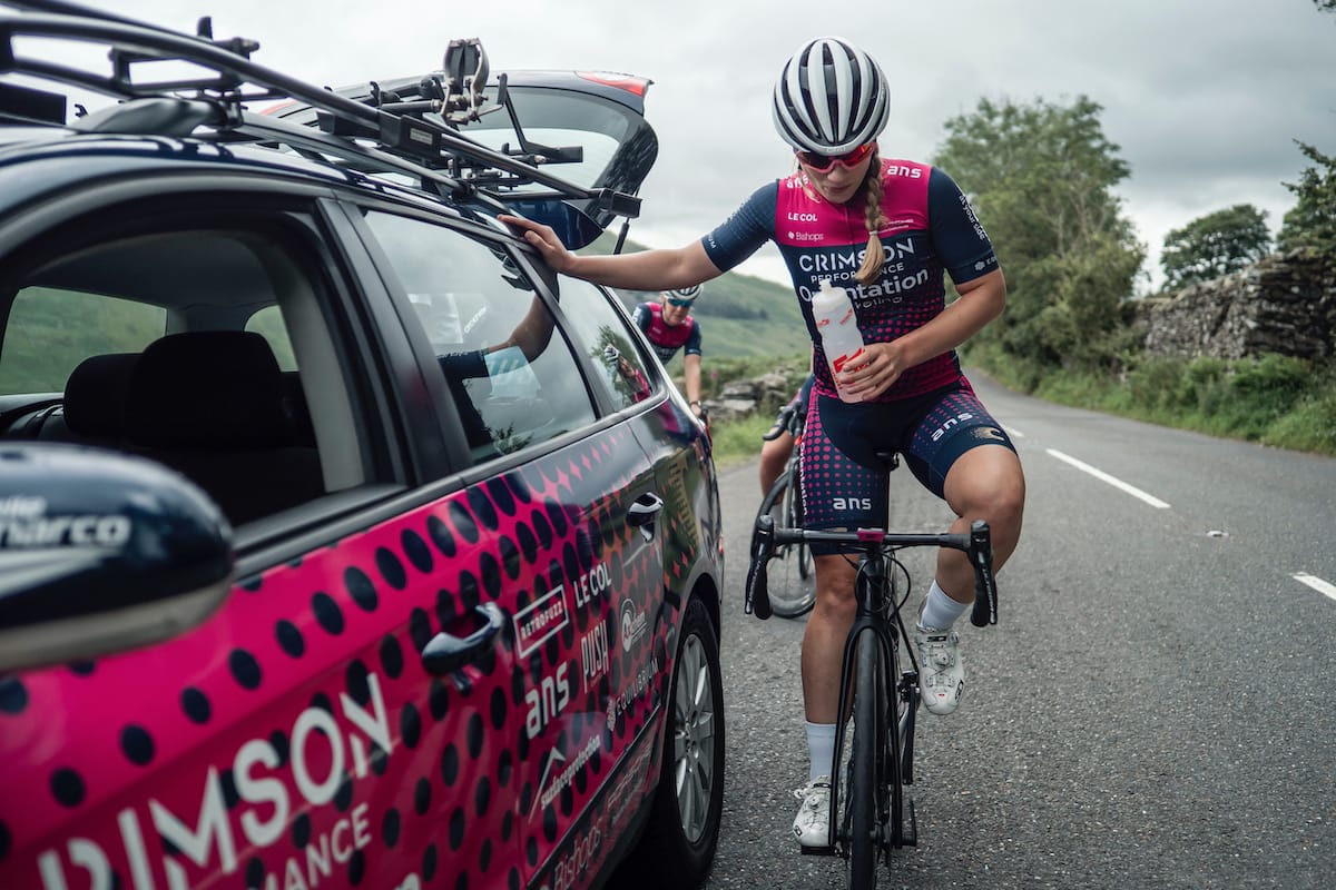 Cyclist Dr Sophie Earl sat on her bike while leaning against a Crimson Performance support vehicle on an empty road in the countryside