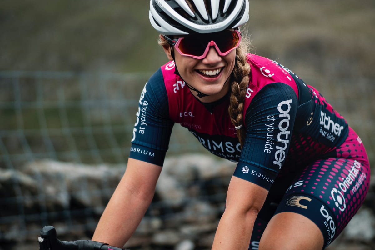 Close-up of cyclist Dr Sophie Earl who's smiling as she rides past with a stone wall and greenery in the background