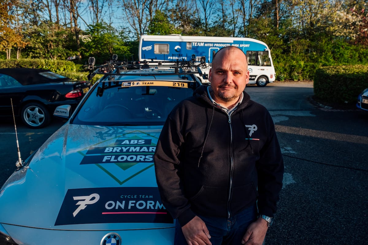 Simon Howes sat on the bonnet of a OnForm support vehicle in a car park with other vehicles in the background