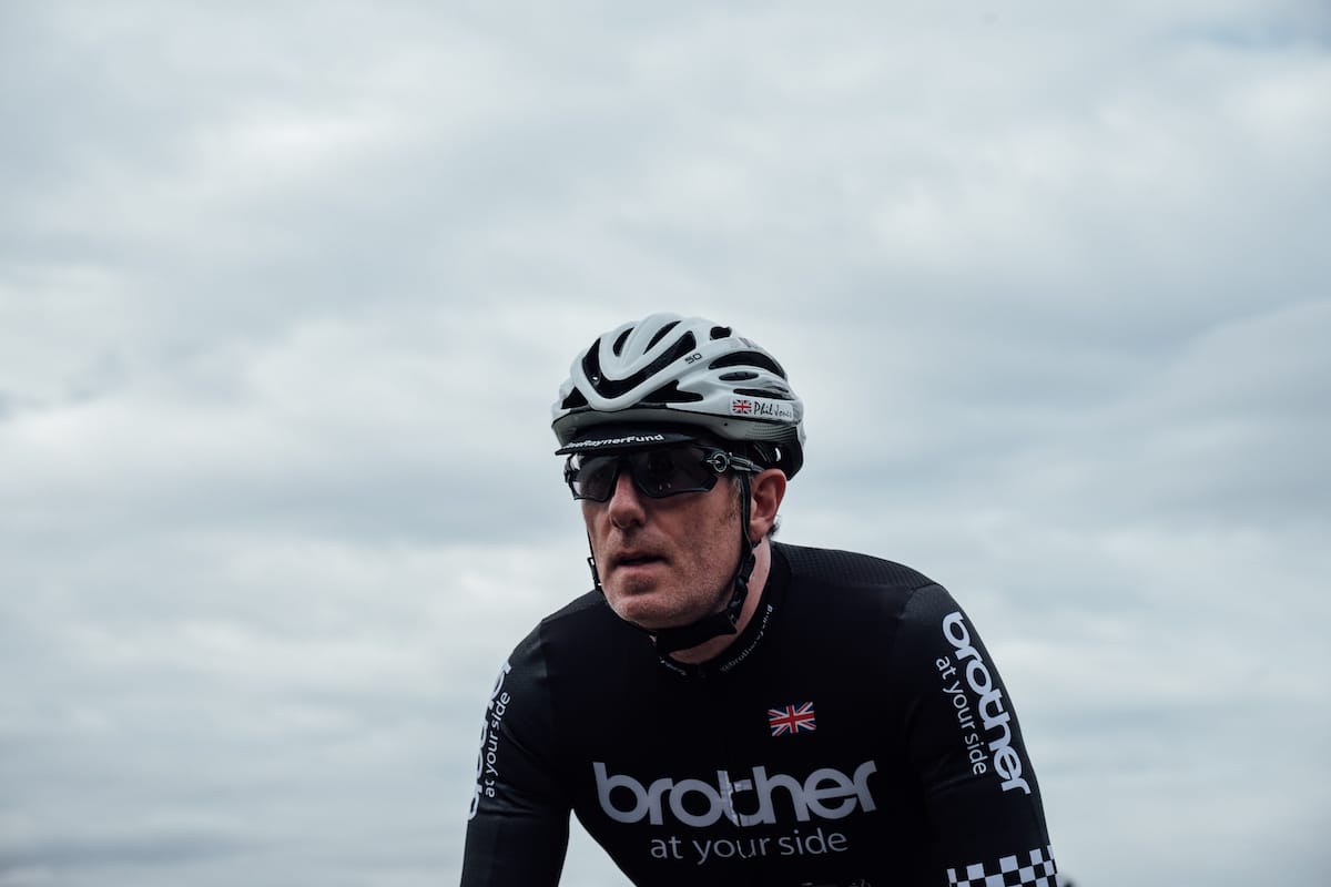 Phil Jones MBE wearing a helmet and Brother cycling team jersey as he rides towards the camera with grey sky in the background