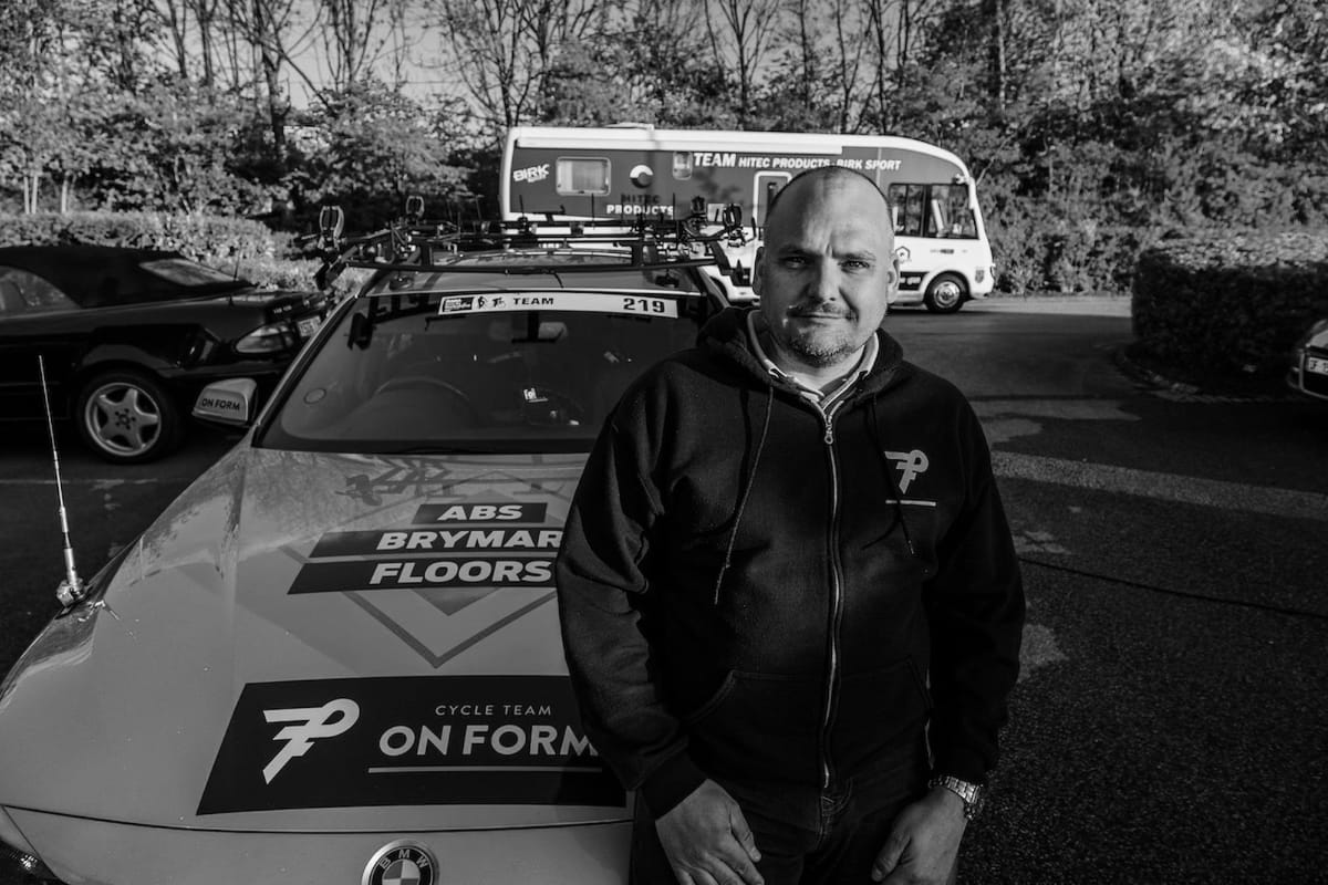 Simon Howes manager of Brother UK-Team OnForm sat on the bonnet of a support vehicle in a car park with other vehicles in the background