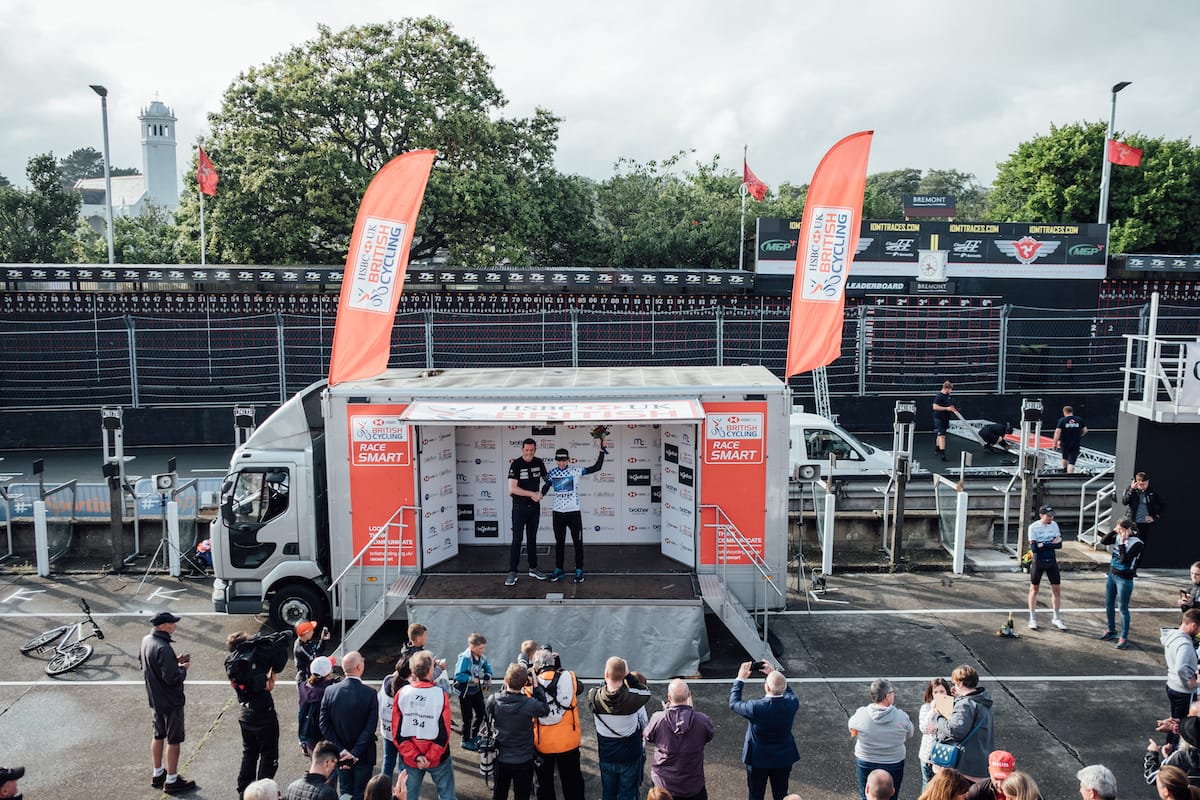 A crowd gathered around a podium built into an open sided truck where a cyclist receives an award at the HSBC National Road Series