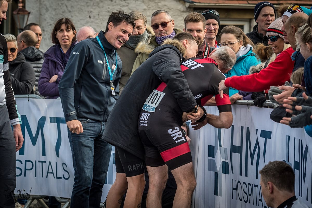 The team assisting an exhausted cyclist who is greeting spectators after a hill climb