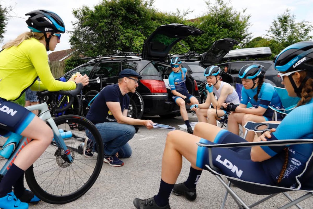 General manager Ian Watson of Team Brother UK-LDN kneeling while giving team talk to cyclists in car park