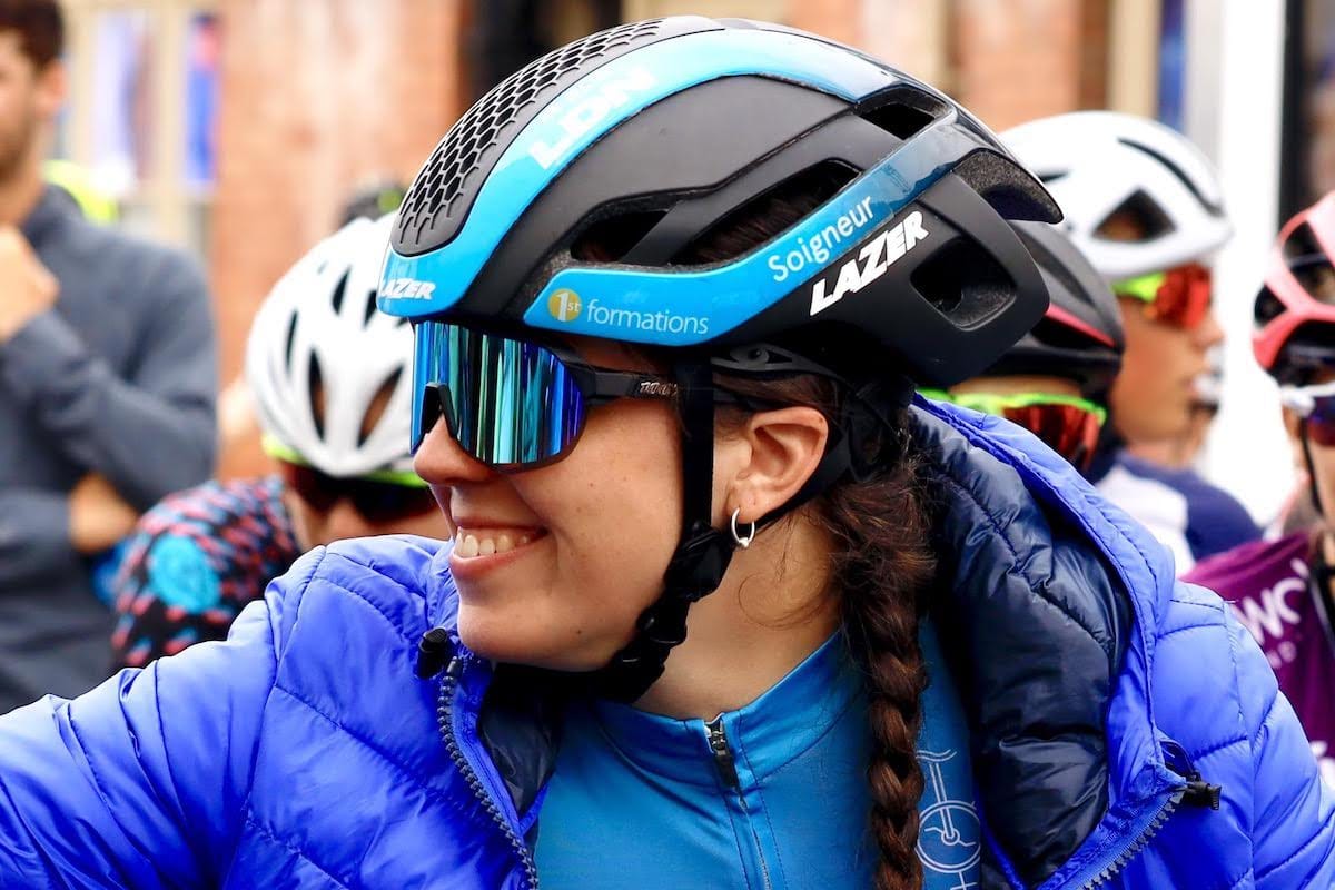 Closeup of Amy Marks smiling to someone out of shot while wearing sunglasses and a helmet with other cyclists in the background