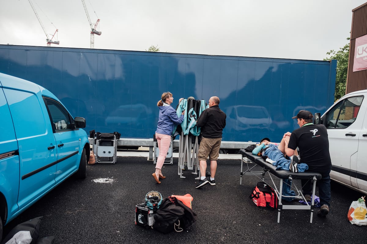 Members of Team OnForm in between two vehicles attending to a folding banner while a physiotherapist is treating a riders leg with a blue container in the background