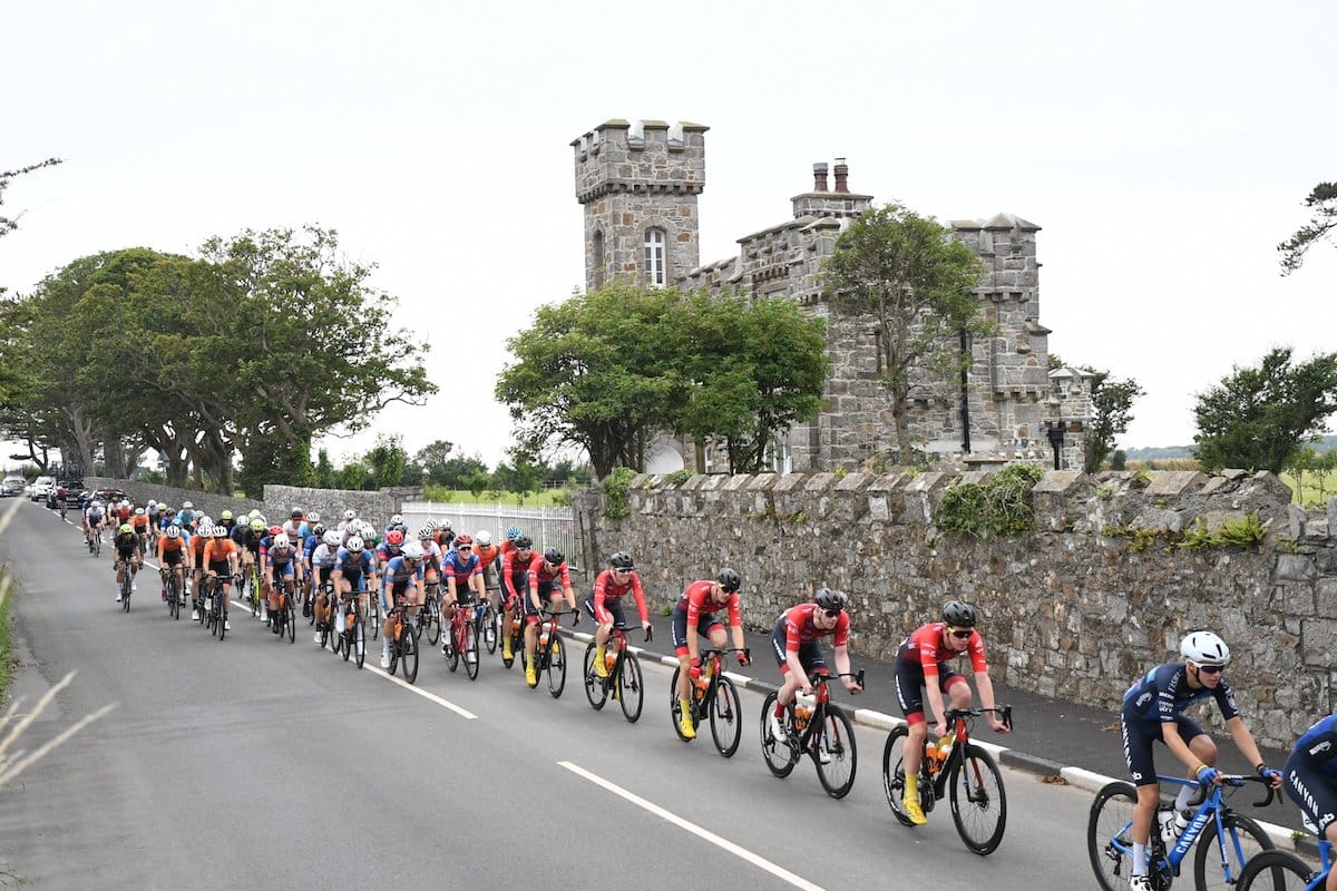 A line of cyclists riding past on a road which has a stone wall and stone building with turrets at the side