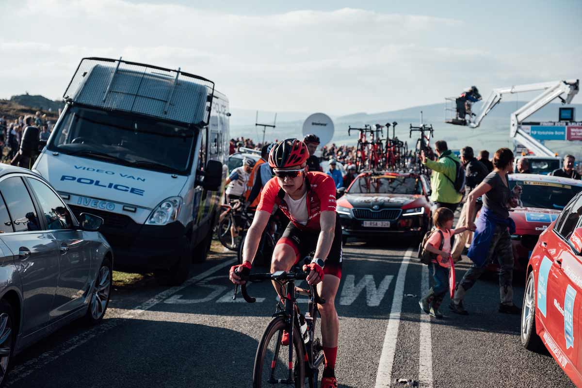 Liam David cycling past parked cars with a support vehicle behind him