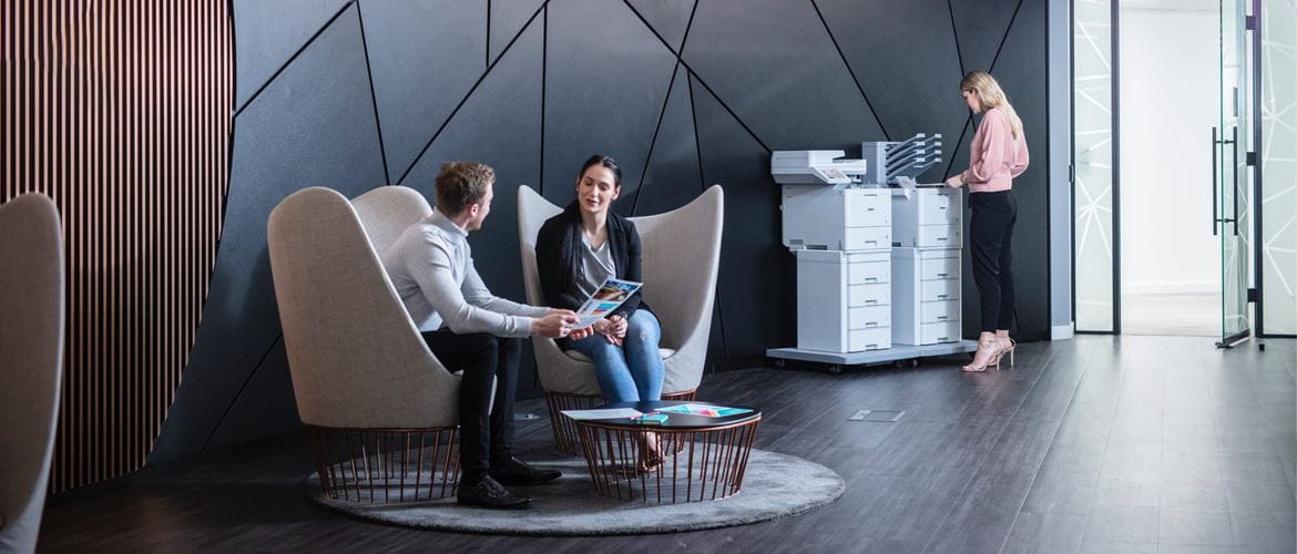 Male and female colleagues having a discussion while sat on high backed chairs in an office lobby with a lady operating two large enterprise printers in the background