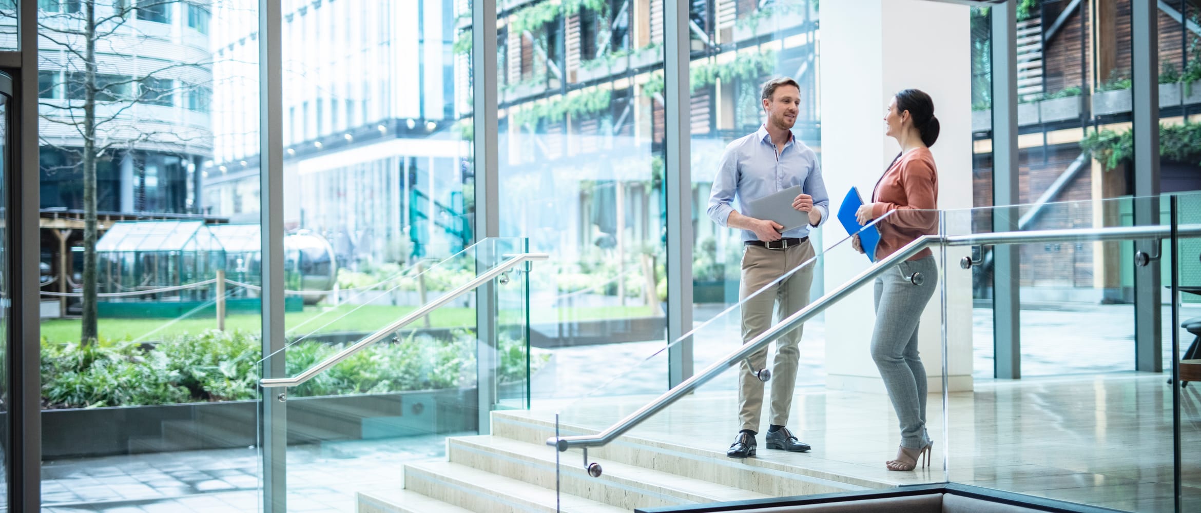 Male and female colleagues chatting at the top of a flight of stair in a glass walled office lobby with a lawned courtyard and other commercial buildings in the background