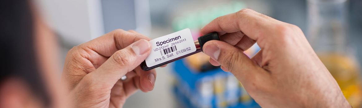 A male healthcare professional in a laboratory applying a barcode label to a test tube containing a blood sample, labelled 'Specimen'