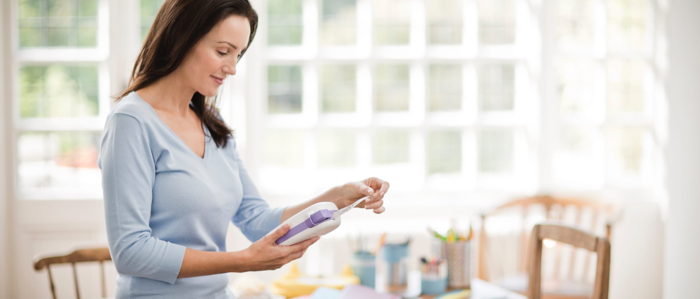 A lady printing a label from a Brother PT-H200 handheld craft label printer while sitting on the edge a table in front of a window in a home environment