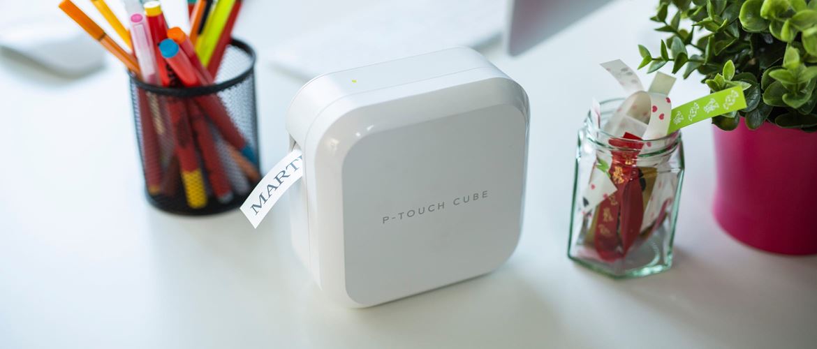 A Brother P-touch CUBE label printer on a white desk surrounded by a pencil jar, ribbon jar, plant and computer