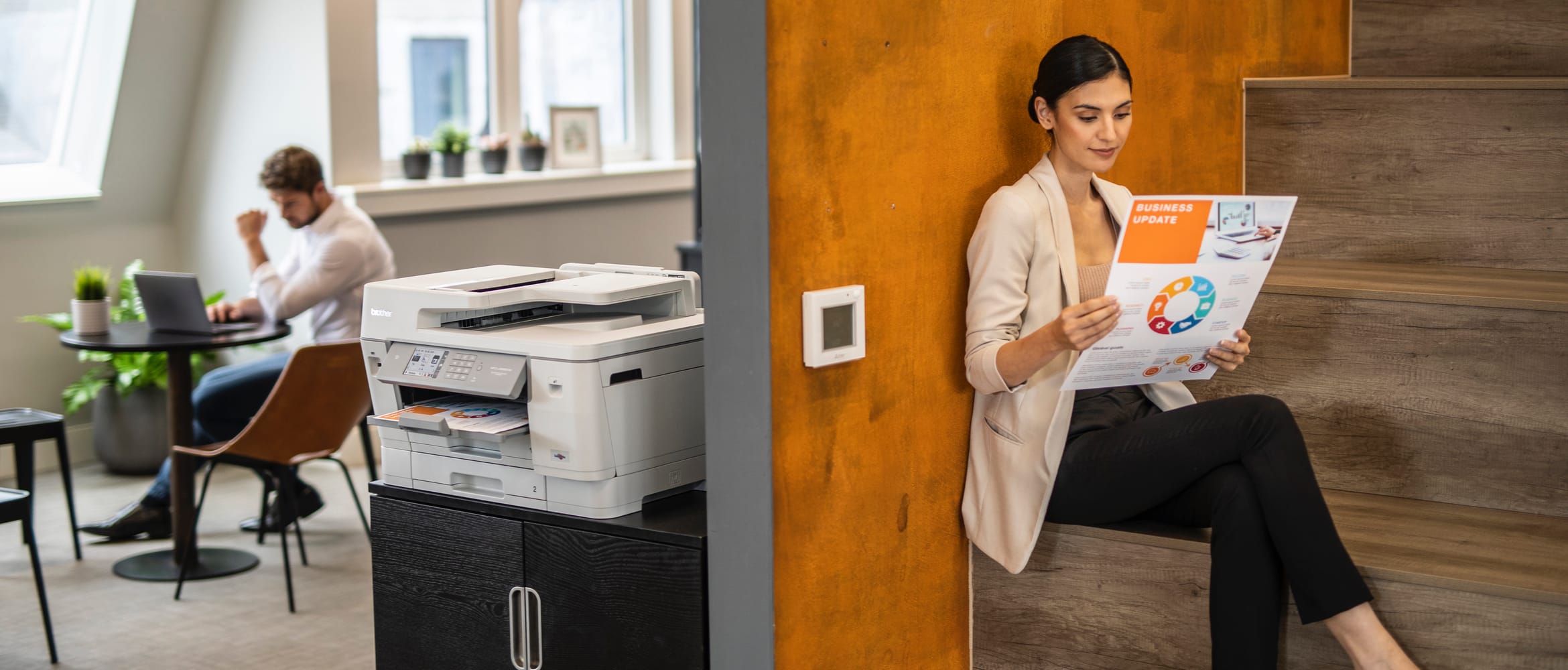 A business woman reading an A3 colour printed document while sat on a staircase behind a Brother printer in an office environment