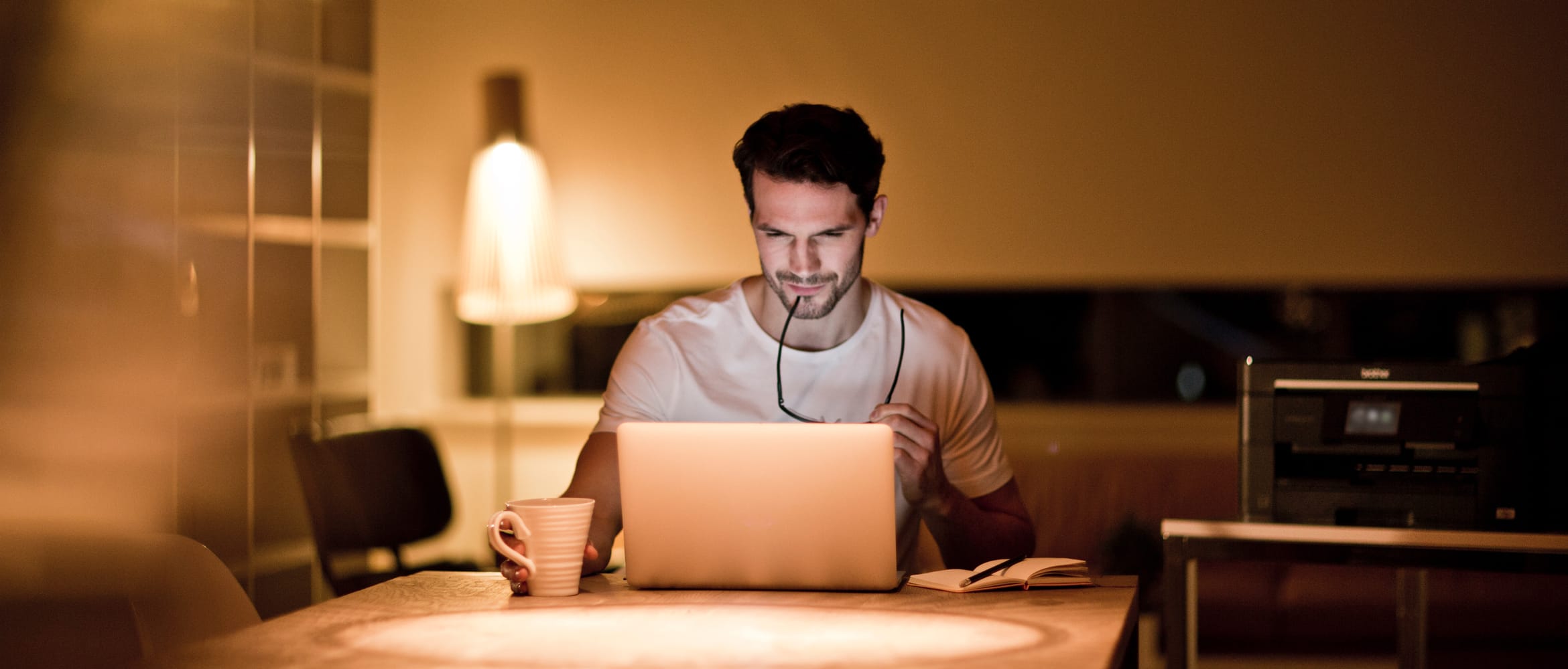 A man reviewing printer offers on a notebook computer while sat at a table in a softly lit room in a home environment