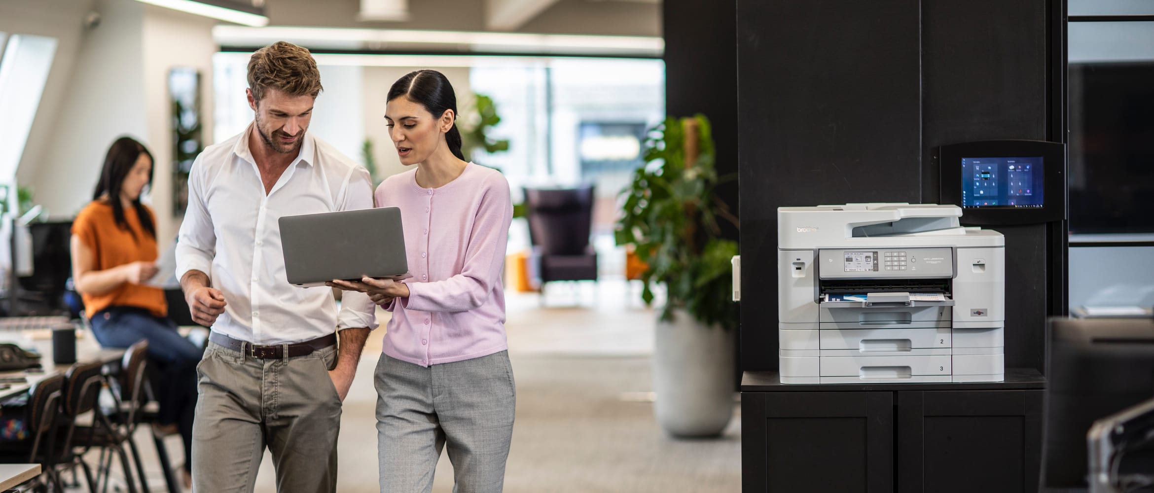 Male and female colleagues printing a document from a notebook computer to a wireless all-in-one printer while walking through an office space