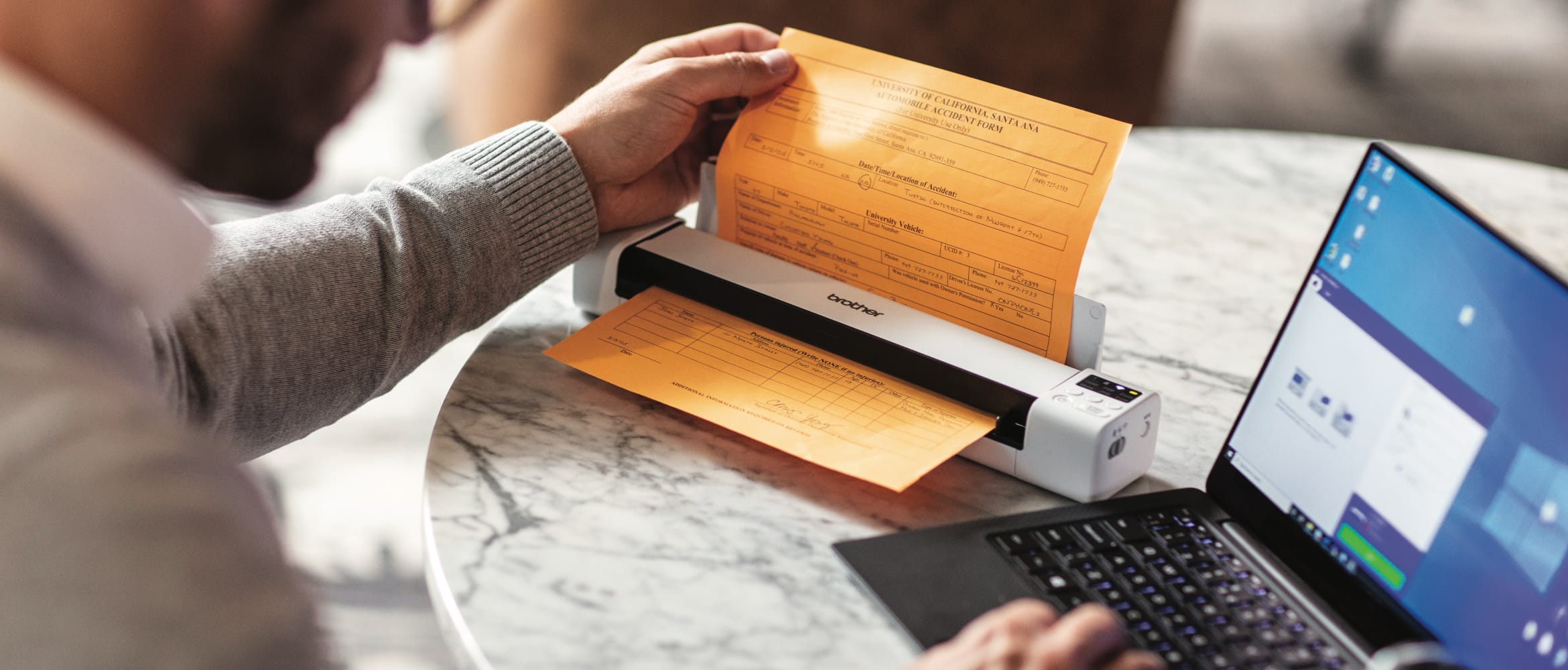 Man using a Brother portable scanner to scan a hand written form to a notebook computer while sat at a marble table