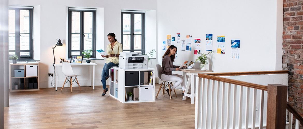 Two ladies, one sat at a desk in front of a notebook computer, the other perched on a shelving unit next to an all-in-one inkjet printer in the centre of a small office