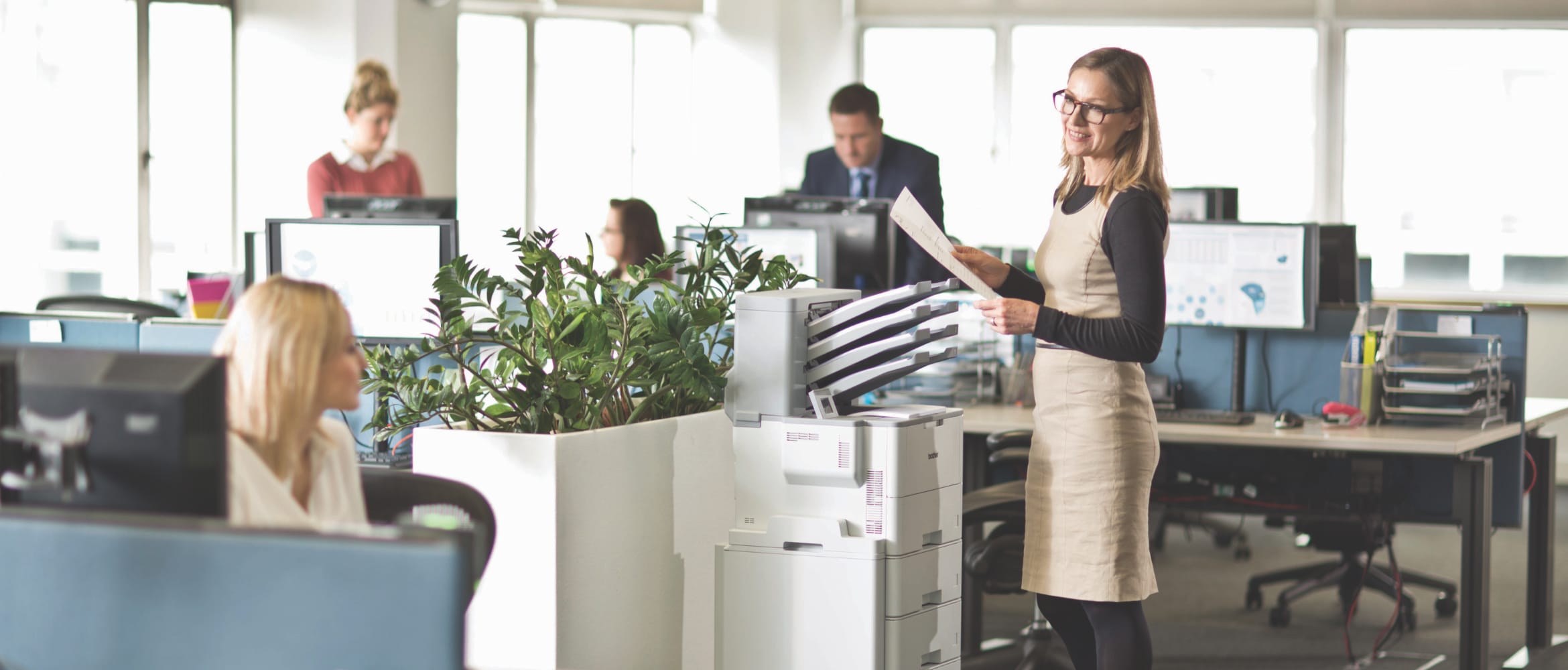 Lady talking to a female colleague while using a Brother HL-L6400DW mono laser workgroup printer in a busy office environment