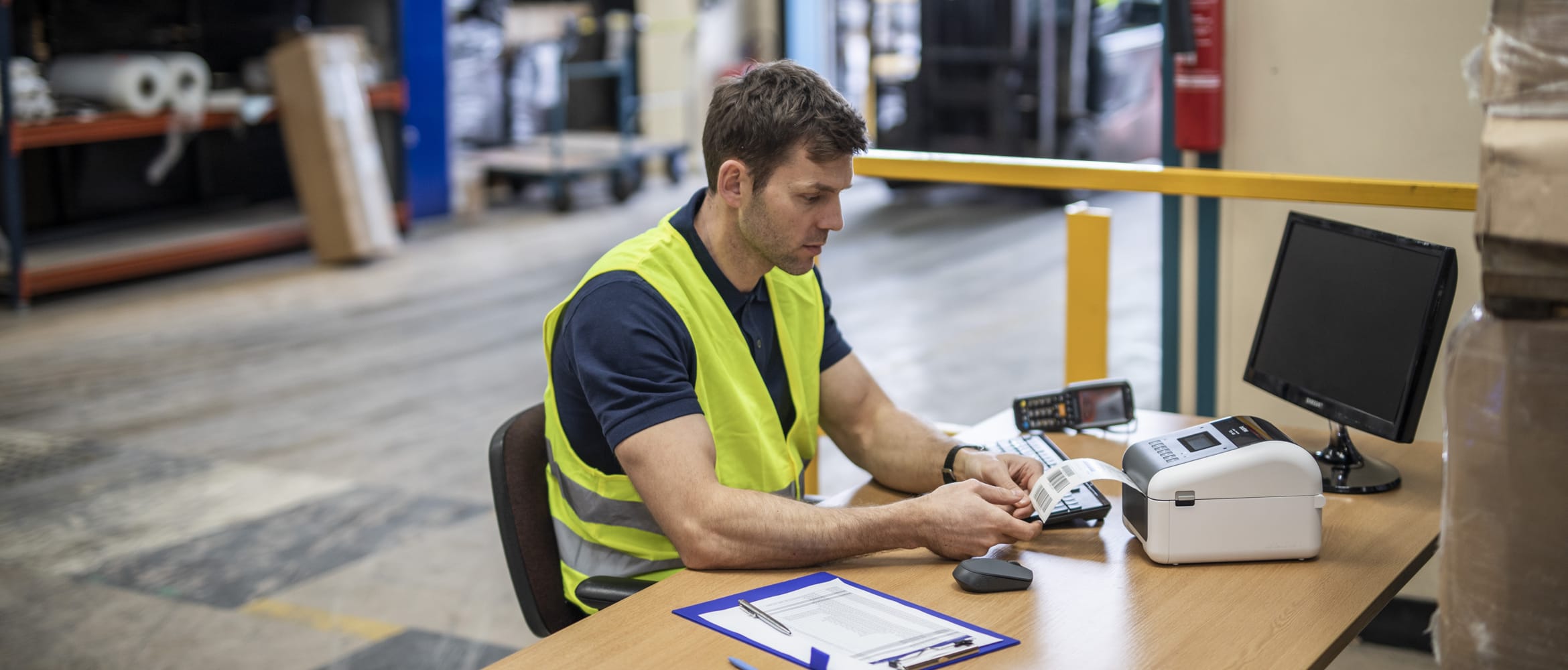 Man with short brown hair wearing yellow hi-vis sat in warehouse at a table, paper, pen, monitor, Brother TD label printer