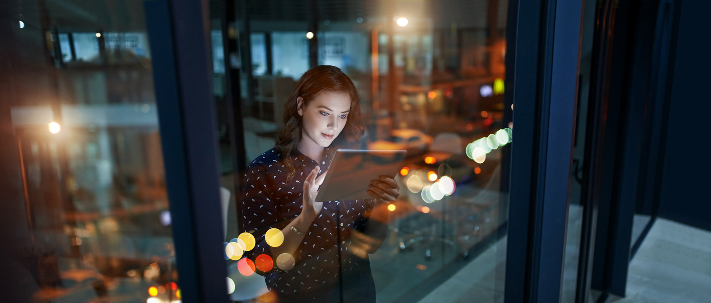 A female office employee is working late in the workplace of the future. She is reviewing a document on her tablet computer while standing next to a window overlooking the city at night. 