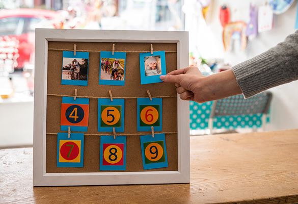 A frame with 3 rows of photos and numbers attached by pegs with a woman's arm outstretched