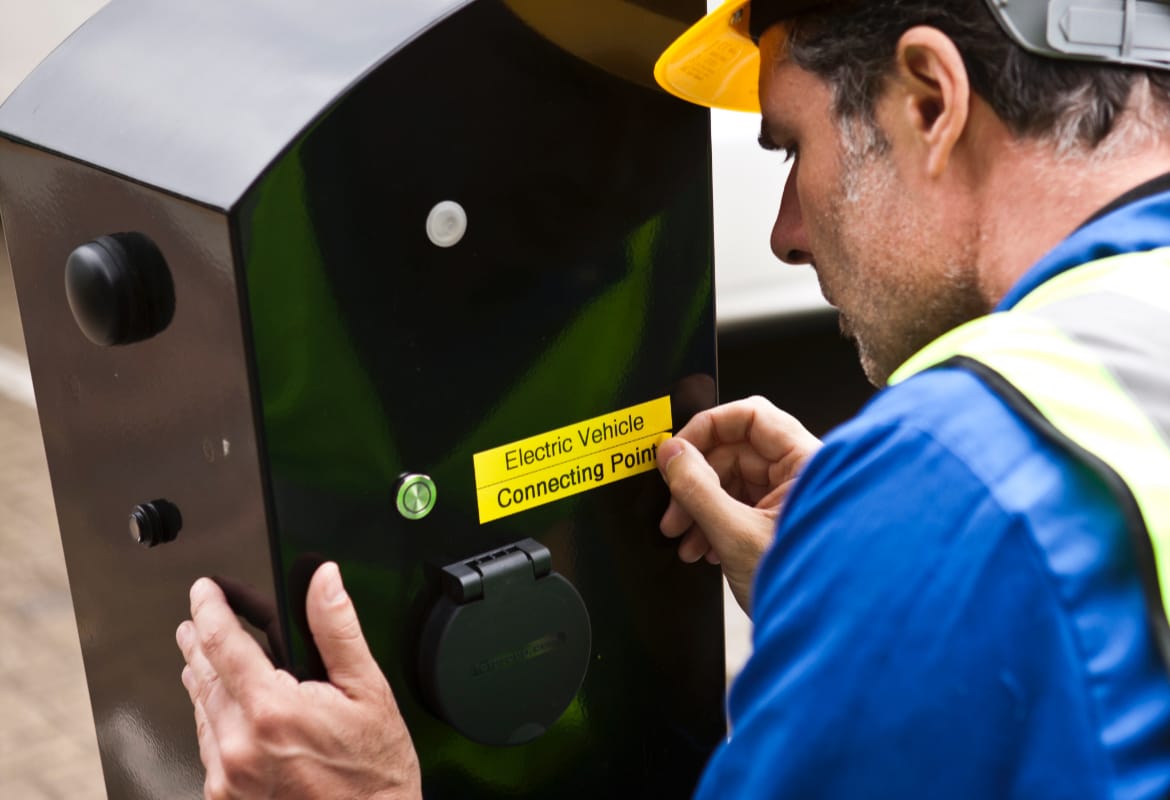 Electrician wearing a hard hat and high visibility vest applying a black on yellow label that reads 'Electric Vehicle Connecting Point' to a black outdoor charger