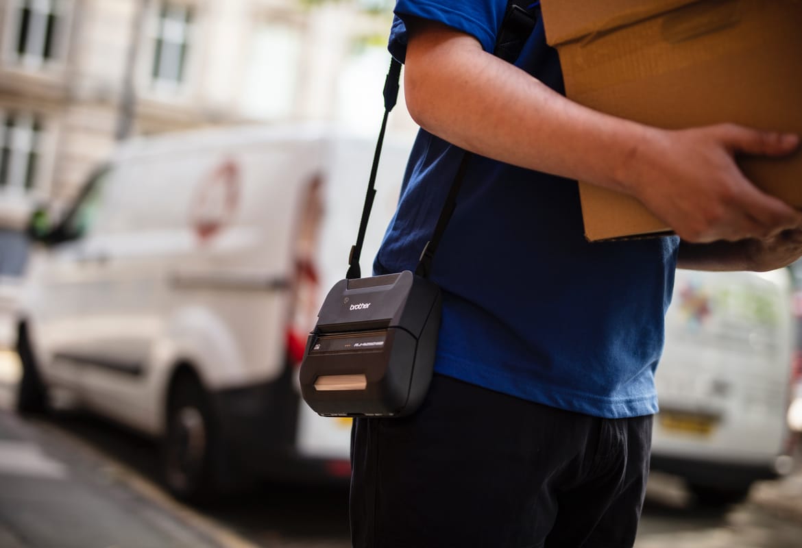 A delivery person carrying cardboard boxes with a Brother RJ Series label printer on a strap over his shoulder with vehicles in the background