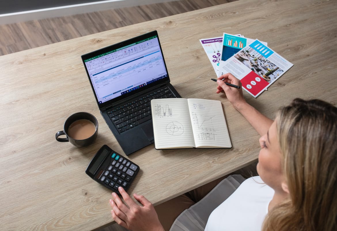 A lady using a calculator, notepad and notebook computer to calculate print costs while sat at a wooden table