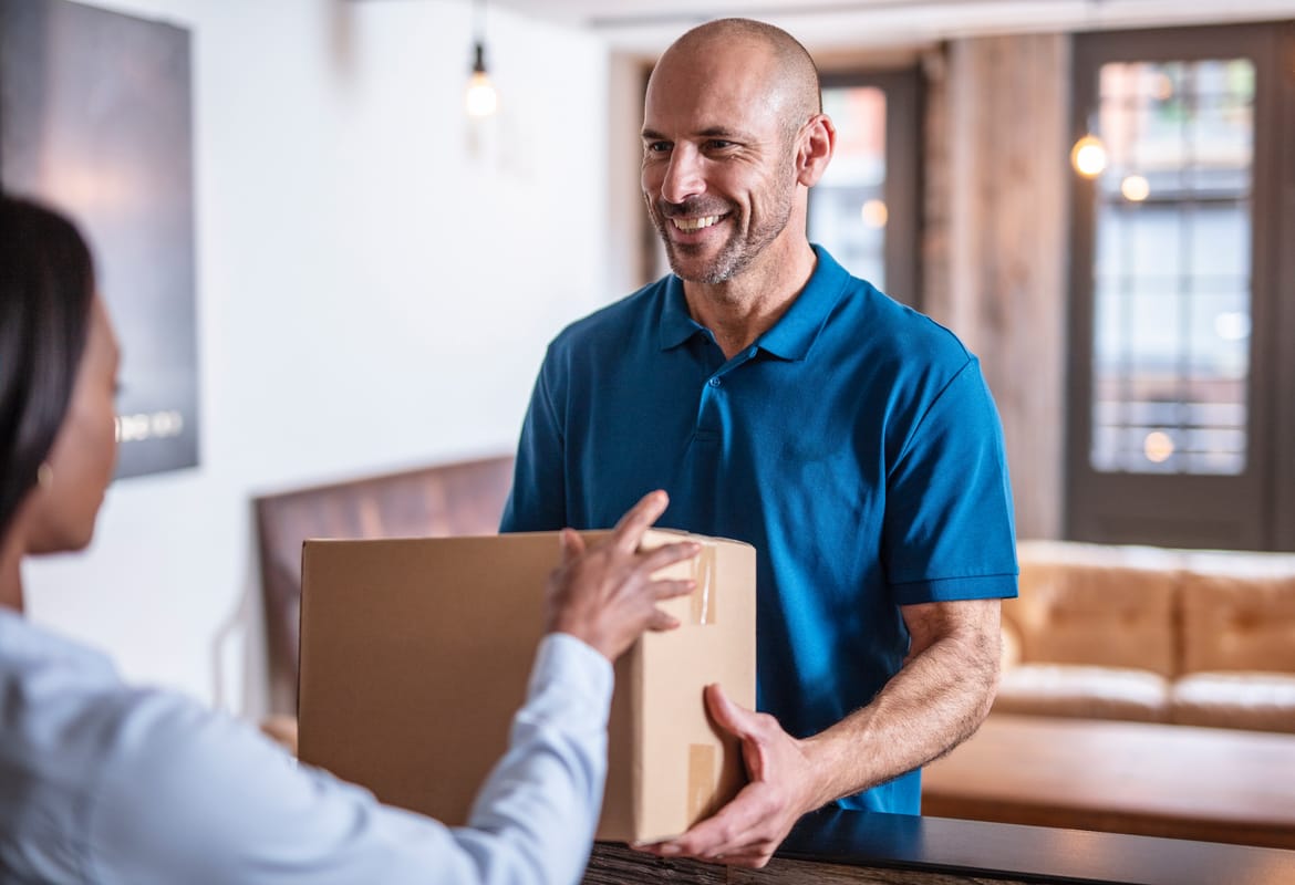 A male delivery person handing a brown cardboard box to a female customer with a coffee table and beige leather sofas in front of glass doors in the background