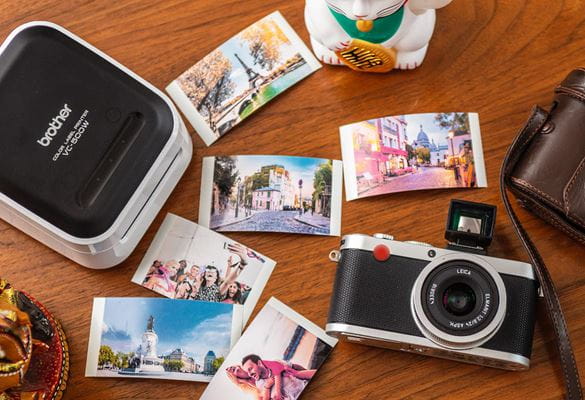 Photographs scattered on a wooden tabletop alongside a Brother VC-500W Design n Craft label printer and a Leica camera