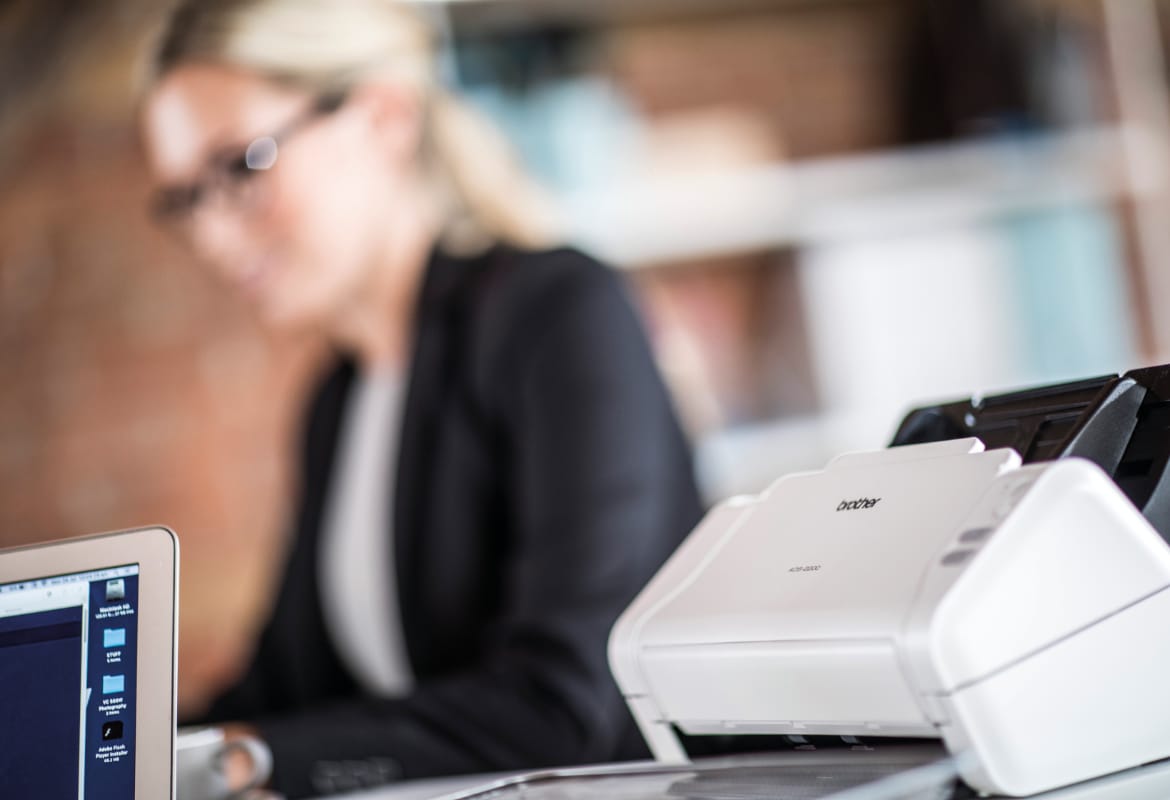 Brother ADS-2200 desktop scanner next to a notebook computer running OCR software with a businesswoman in the background