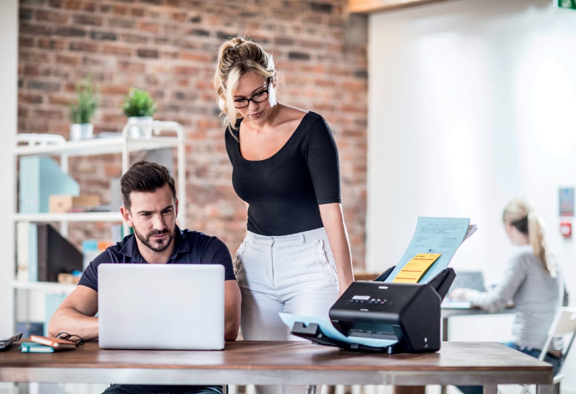 A man using a notebook computer to scan documents from a Brother ADS-3000N sheetfed scanner while sat at a desk with a female colleague standing next to him