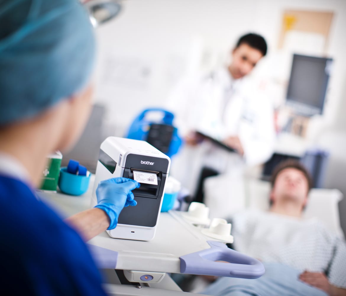 A female doctor removing a barcode ID label from a Brother label printer with a male doctor standing by a patient's bedside in the background