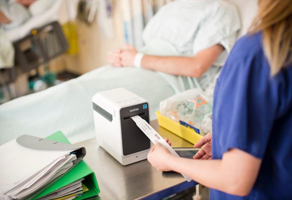 Healthcare professional removing an identification wristband from a label printer with a patient sat upright in a hospital bed in the background