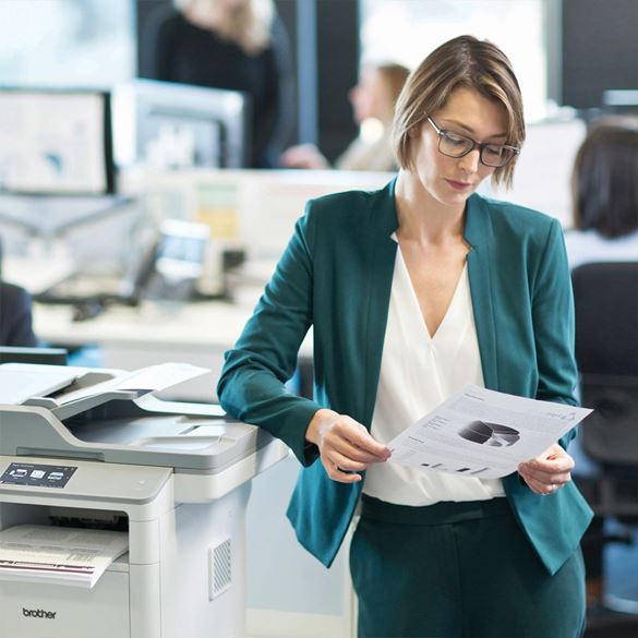 A woman in a teal suit standing next to a Brother printer and reviewing some printed paper
