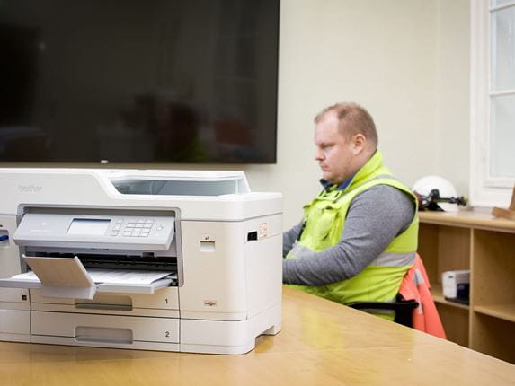 An Alasen Rakennus employee wearing a high visibility vest while sat in an office with a compact Brother A3 inkjet printer with cloud connectivity on a desk in the foreground