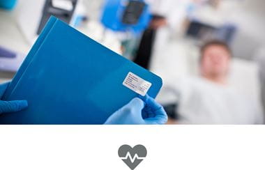 A medical professional applying a label to a blue folder with a patient lying on a hospital bed in the background