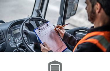 A male HGV driver wearing a high visibility vest writing a report on a clipboard in the drivers seat of his cab