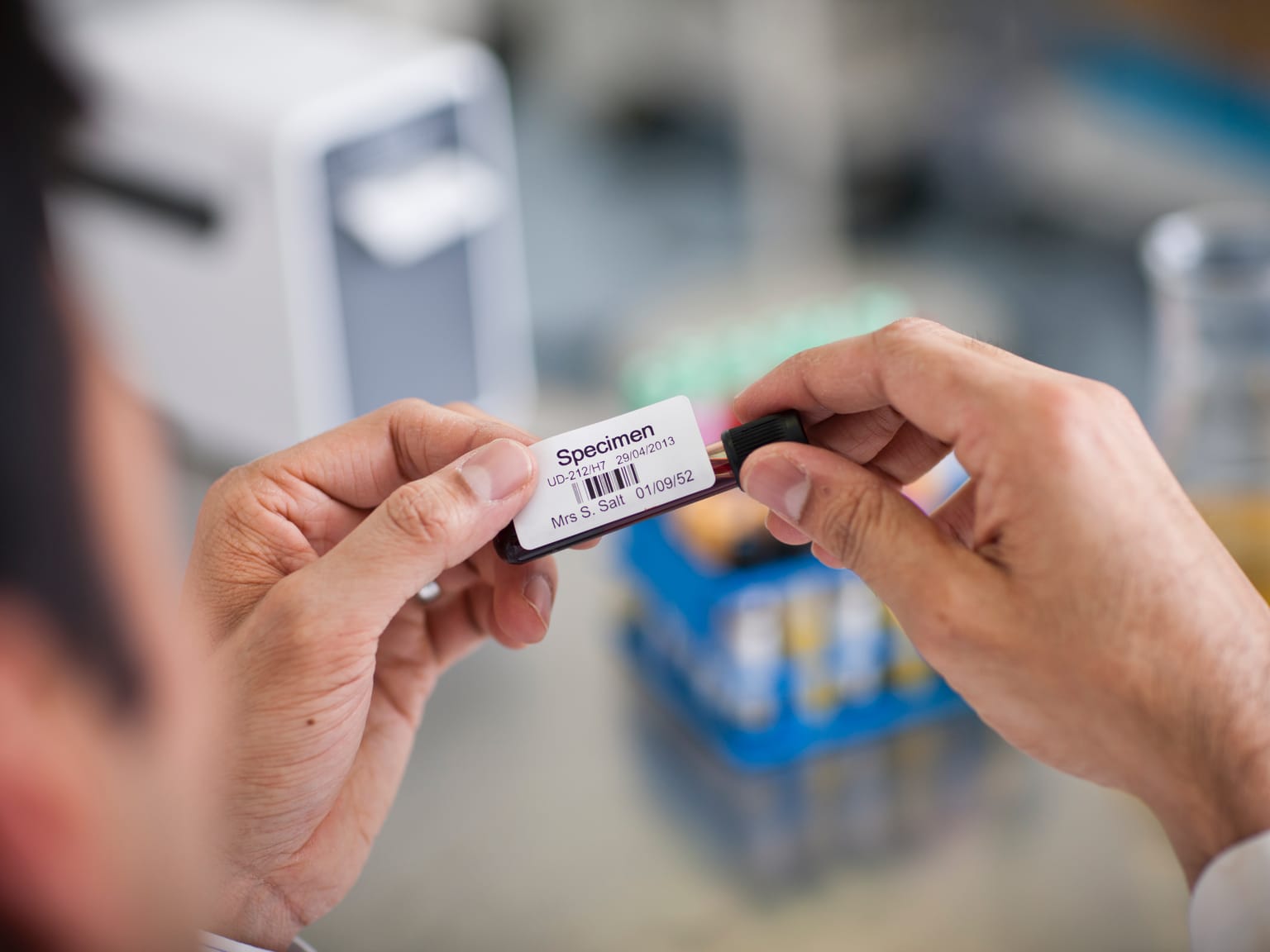 A male healthcare professional in a laboratory applying a barcode label to a test tube containing a blood sample, labelled 'Specimen'
