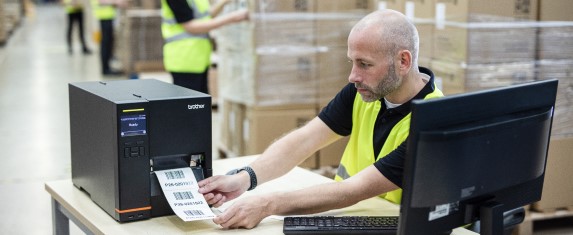 A man in a warehouse wearing a high-vis jacket who is taking a freshly printed label out of a Brother TJ printer