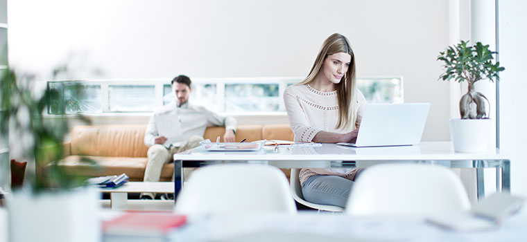 Female sat at desk with laptop, man sat at back on orange sofa, plants, notebooks, white chairs, window