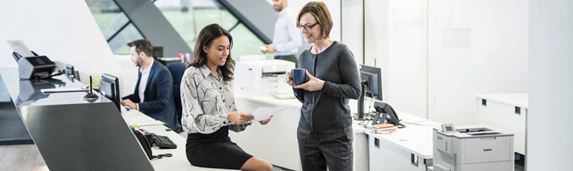 Two ladies in an office looking at a piece of paper and chatting with colleagues and a printer in the background
