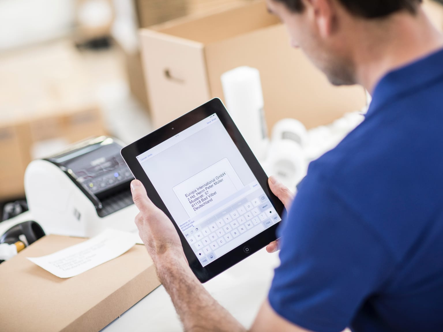 A man using Brother's iPrint&Label app on a tablet device to print a return shipping label on a Brother label printer, with packing boxes in the background.