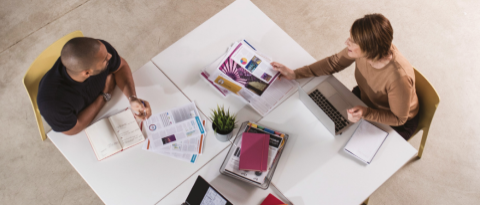 Overhead view of a man and a woman sat at a table and discussing printed work documents 