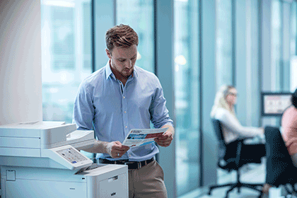 Man next to printer holding colour document, women sat at desks, monitor, chair