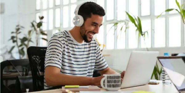 A man wearing headphones working on his laptop and smiling at the screen
