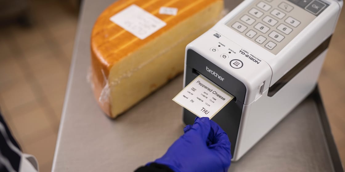 A person out of shot wearing a blue latex glove removing a food label from a Brother TD-2120N industrial label printer, next to a block of cheese in a commercial kitchen environment
